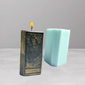 Può includere: Una candela rettangolare accesa con un design in rilievo color bronzo raffigurante una ruota e le parole "WHEEL of FORTUNE". Un blocco rettangolare azzurro è accanto alla candela. La fiamma della candela è gialla e arancione.