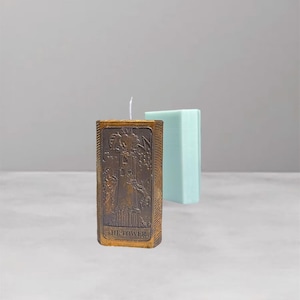 Può includere: Una candela quadrata color oro con un rilievo dettagliato di una torre, etichettata "THE TOWER". Dietro c'è una candela rettangolare azzurra. Le candele sono su uno sfondo neutro.