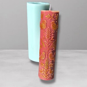 Puede incluir: Una vela de pilar de color rojo anaranjado adornada con patrones en relieve dorados de p&aacute;jaros y flores. Un molde cil&iacute;ndrico azul claro est&aacute; posicionado detr&aacute;s de la vela. El dise&ntilde;o de la vela presenta motivos bot&aacute;nicos y aviares detallados.