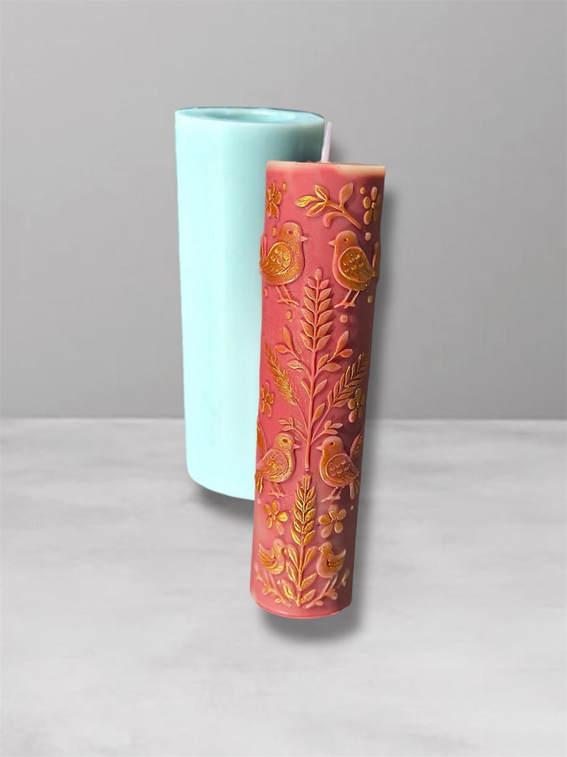 Puede incluir: Una vela de pilar de color rojo anaranjado adornada con patrones en relieve dorados de p&aacute;jaros y flores. Un molde cil&iacute;ndrico azul claro est&aacute; posicionado detr&aacute;s de la vela. El dise&ntilde;o de la vela presenta motivos bot&aacute;nicos y aviares detallados.