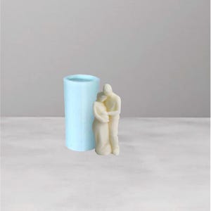 May include: A light blue cylindrical candle mold next to a cream-colored sculpture of two figures embracing. The figures are stylized, with smooth, rounded forms. The mold is made of a translucent material.