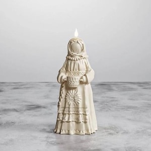 Könnte beinhalten: Eine cremefarbene Kerze in Form einer bekleideten Figur, die einen Korb hält. Die Kerze hat oben ein flammenförmiges Licht. Die Figur hat detaillierte Schnitzereien, darunter ein Sonnendesign auf dem Rock. Die Kerze steht auf einer grauen Oberfläche.