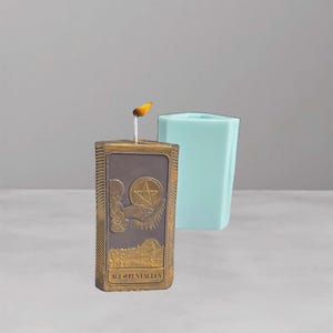 Puede incluir: Una vela encendida con un diseño rectangular dorado que presenta la carta del tarot As de Oros. La vela tiene un fondo marrón y un borde dorado. Un objeto hexagonal azul claro está en el fondo.