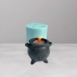 May include: A black cauldron-shaped candle with a burning flame, featuring an eye symbol and text. A light blue cylindrical candle is in the background. The candle is on a white surface.