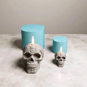 May include: Two gray skull-shaped candles with lit flames, one large and one small, sit on a marble surface. The skulls have detailed carvings. Two light blue cylindrical objects are in the background.