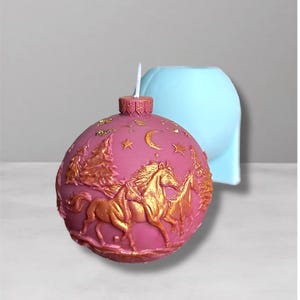 Könnte beinhalten: Eine rosa weihnachtskugelförmige Kerze mit einem goldenen Pferde- und Baumdesign. Die Kerze hat einen weißen Docht und eine dekorative goldene Oberseite. Eine hellblaue Form befindet sich im Hintergrund.