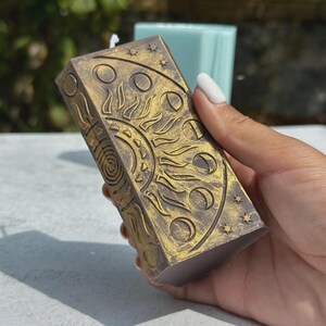 Puede incluir: Una vela rectangular con un dise&ntilde;o celestial. La vela es de color p&uacute;rpura apagado con detalles dorados. El dise&ntilde;o incluye un sol, fases lunares y estrellas. La vela est&aacute; sostenida en una mano.