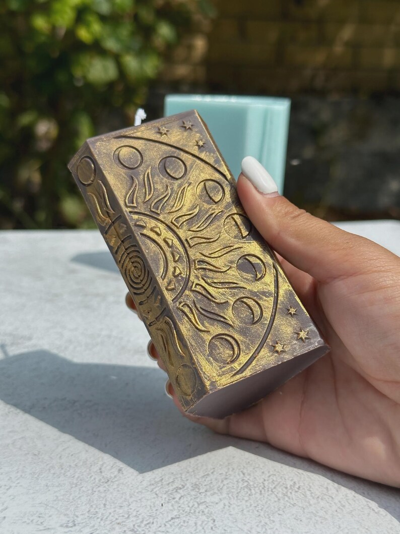 Puede incluir: Una vela rectangular con un dise&ntilde;o celestial. La vela es de color p&uacute;rpura apagado con detalles dorados. El dise&ntilde;o incluye un sol, fases lunares y estrellas. La vela est&aacute; sostenida en una mano.