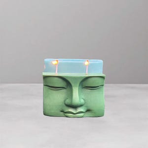 May include: A green Buddha face candle with a light blue top section. Two lit wicks are visible. The candle has a serene expression and is set against a gray background.