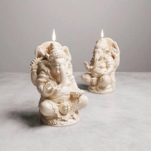 May include: Two ivory-colored Ganesh candles are depicted, each with a lit wick. The candles are intricately sculpted in the likeness of the Hindu deity Ganesh, seated in a meditative pose. The background is a neutral gray, highlighting the detailed craftsmanship.