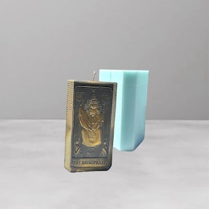 Può includere: Una candela rettangolare con un design oro e nero con la scritta "THE HIEROPHANT" e uno stampo azzurro a forma geometrica. La candela ha uno stoppino ed è probabilmente fatta a mano. Lo sfondo è grigio chiaro.