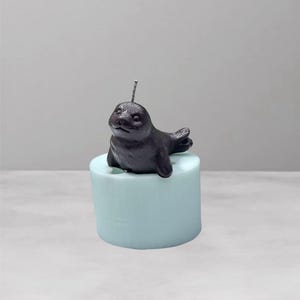 May include: A dark gray seal candle with a wick, resting on a light blue cylindrical base. The candle has a detailed seal figure, perfect for home decor or as a unique gift. The candle is a charming addition to any room.