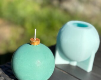 May include: A spherical teal candle with a gold-colored cap and a white wick, positioned next to a light blue silicone mold shaped like a sphere with a flat base, designed for casting round objects in a crafting environment.