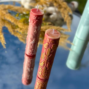 Puede incluir: Dos velas de pilar decorativas con intrincados dise&ntilde;os florales y de p&aacute;jaros. Una vela es de color rosa claro con detalles blancos, y la otra es de color rosa m&aacute;s oscuro con detalles dorados. Las velas son cil&iacute;ndricas y tienen mechas blancas.