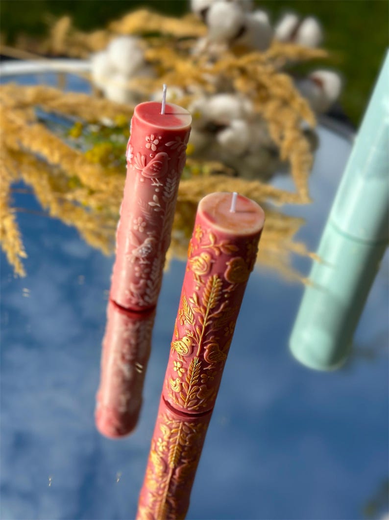 Puede incluir: Dos velas de pilar decorativas con intrincados dise&ntilde;os florales y de p&aacute;jaros. Una vela es de color rosa claro con detalles blancos, y la otra es de color rosa m&aacute;s oscuro con detalles dorados. Las velas son cil&iacute;ndricas y tienen mechas blancas.
