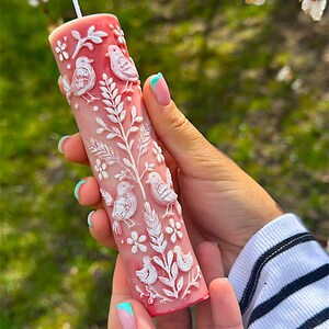 Puede incluir: Una vela de pilar rosa con tallas en relieve blancas de p&aacute;jaros, flores y follaje. La vela es cil&iacute;ndrica y tiene una mecha blanca. El dise&ntilde;o es detallado y la vela se sostiene en una mano.