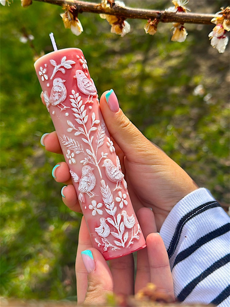 Puede incluir: Una vela de pilar rosa con tallas en relieve blancas de p&aacute;jaros, flores y follaje. La vela es cil&iacute;ndrica y tiene una mecha blanca. El dise&ntilde;o es detallado y la vela se sostiene en una mano.