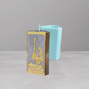 Puede incluir: Una vela rectangular con un diseño de carta del tarot, con la carta "As de Bastos". La vela tiene un fondo gris con detalles dorados y un borde marrón. Un molde geométrico azul claro está en el fondo.