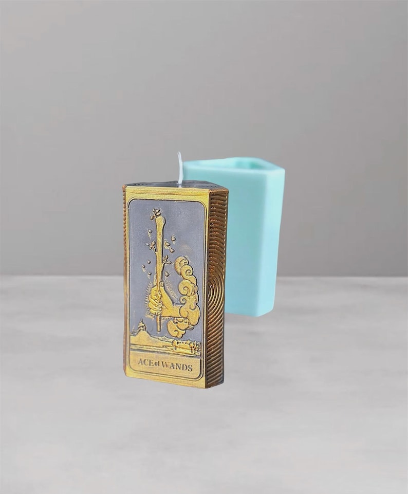 Puede incluir: Una vela rectangular con un dise&ntilde;o de carta del tarot, con la carta "As de Bastos". La vela tiene un fondo gris con detalles dorados y un borde marr&oacute;n. Un molde geom&eacute;trico azul claro est&aacute; en el fondo.