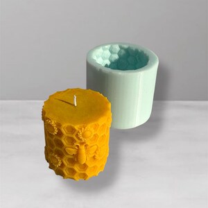 May include: A yellow, cylindrical candle with a honeycomb and bee design, next to a light blue silicone mold. The candle has a detailed texture and a white wick. The mold has a honeycomb pattern inside.