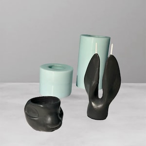 Pode incluir: Uma coleção de velas e moldes de velas feitos à mão. As velas incluem uma vela preta em forma de coelho, uma pequena vela preta em forma de rosto e uma vela alta azul clara. Também estão presentes dois moldes de velas azuis claras.
