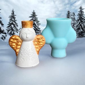 May include: A white angel figurine with gold-colored wings and a gold crown, next to a light blue, abstract-shaped object. The angel has a snowflake design on its dress. The background features a snowy landscape with evergreen trees.