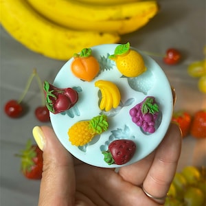 May include: A light blue silicone mold filled with miniature fruit shapes, including a peach, lemon, banana, pineapple, strawberry, grapes, and cherries. Yellow bananas are in the background.
