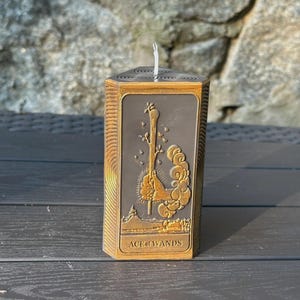Puede incluir: Una vela hexagonal con un dise&ntilde;o detallado de la carta del tarot As de Bastos. La vela tiene un marco de color bronce y un centro gris oscuro con la imagen de la carta. El texto "ACE of WANDS" est&aacute; en la parte inferior.