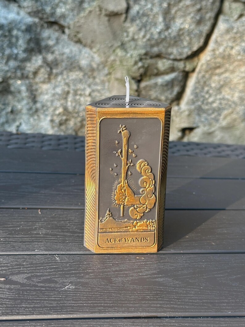 Puede incluir: Una vela hexagonal con un dise&ntilde;o detallado de la carta del tarot As de Bastos. La vela tiene un marco de color bronce y un centro gris oscuro con la imagen de la carta. El texto "ACE of WANDS" est&aacute; en la parte inferior.