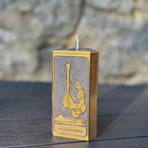 Puede incluir: Una vela rectangular con un dise&ntilde;o dorado y gris que presenta la carta del tarot As de Bastos. La vela tiene una mecha blanca y est&aacute; colocada sobre una superficie oscura. El texto "ACE of WANDS" es visible.