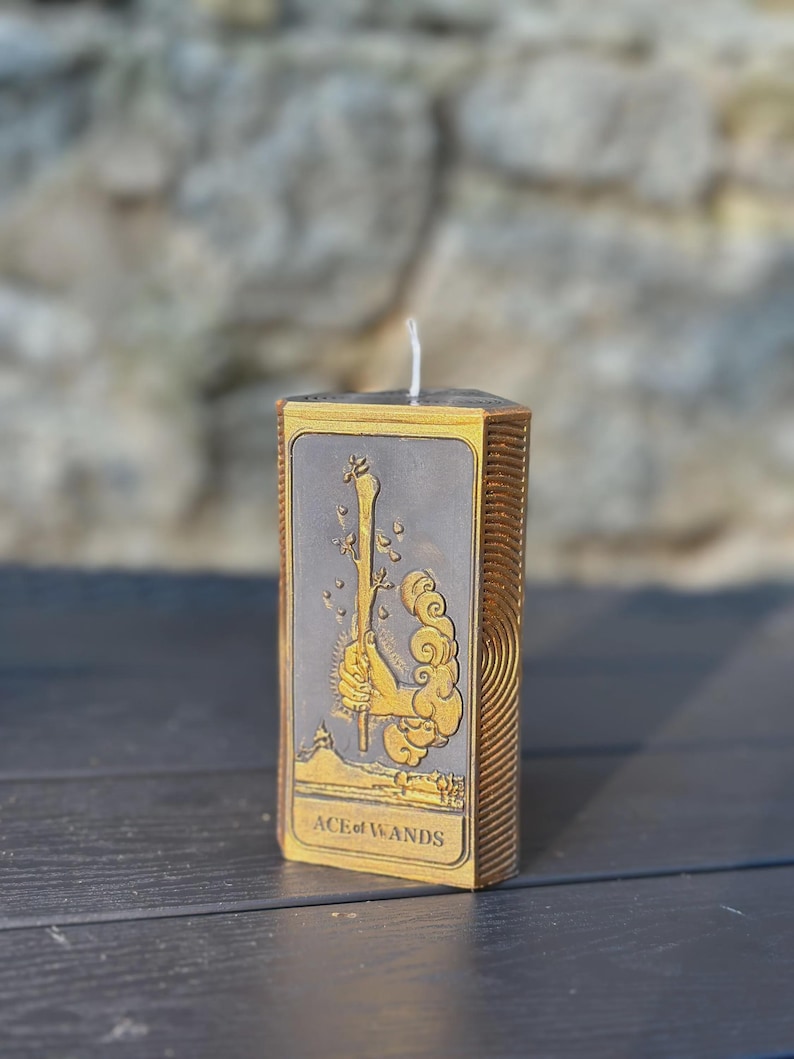 Puede incluir: Una vela rectangular con un dise&ntilde;o dorado y gris que presenta la carta del tarot As de Bastos. La vela tiene una mecha blanca y est&aacute; colocada sobre una superficie oscura. El texto "ACE of WANDS" es visible.