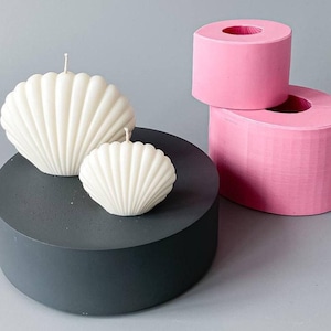 May include: Two seashell-shaped candles with wicks sit on a dark gray cylindrical platform. A larger candle is next to a smaller one. Two pink cylindrical molds are in the background. The candles are white.