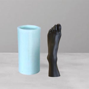 以下が含まれることがあります： 水色の円筒形のキャンドルホルダーと、黒い足の形をした彫刻。キャンドルホルダーは無地で不透明な色です。足の彫刻は、人間の足の詳細な表現です。