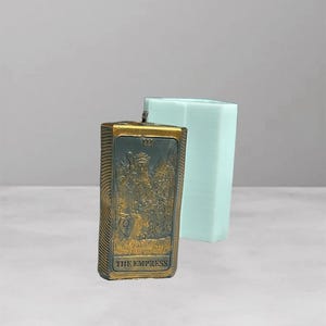 Puede incluir: Una vela rectangular con un diseño dorado y bronce que presenta el texto "THE EMPRESS" y una ilustración detallada. Un molde hexagonal azul claro está detrás de la vela.