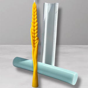 Puede incluir: Una vela amarilla encendida con forma de espiga de trigo, con un molde azul y un cilindro azul. La vela tiene una llama en la parte superior. La vela es amarilla y tiene una textura detallada. El molde es transparente y tiene forma rectangular. El cilindro es azul.
