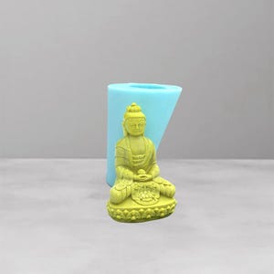 May include: A yellow-green Buddha statue in a seated meditation pose. The statue is detailed with intricate carvings and sits on a decorative base. A light blue mold is behind the statue. The background is a light gray.