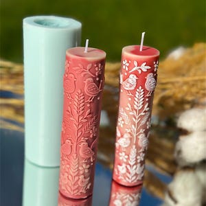 Puede incluir: Dos velas de pilar rojas con dise&ntilde;os de p&aacute;jaros y flores blancos. Un molde cil&iacute;ndrico azul claro para velas est&aacute; en el fondo. Las velas tienen una superficie lisa y reflectante y una mecha blanca.
