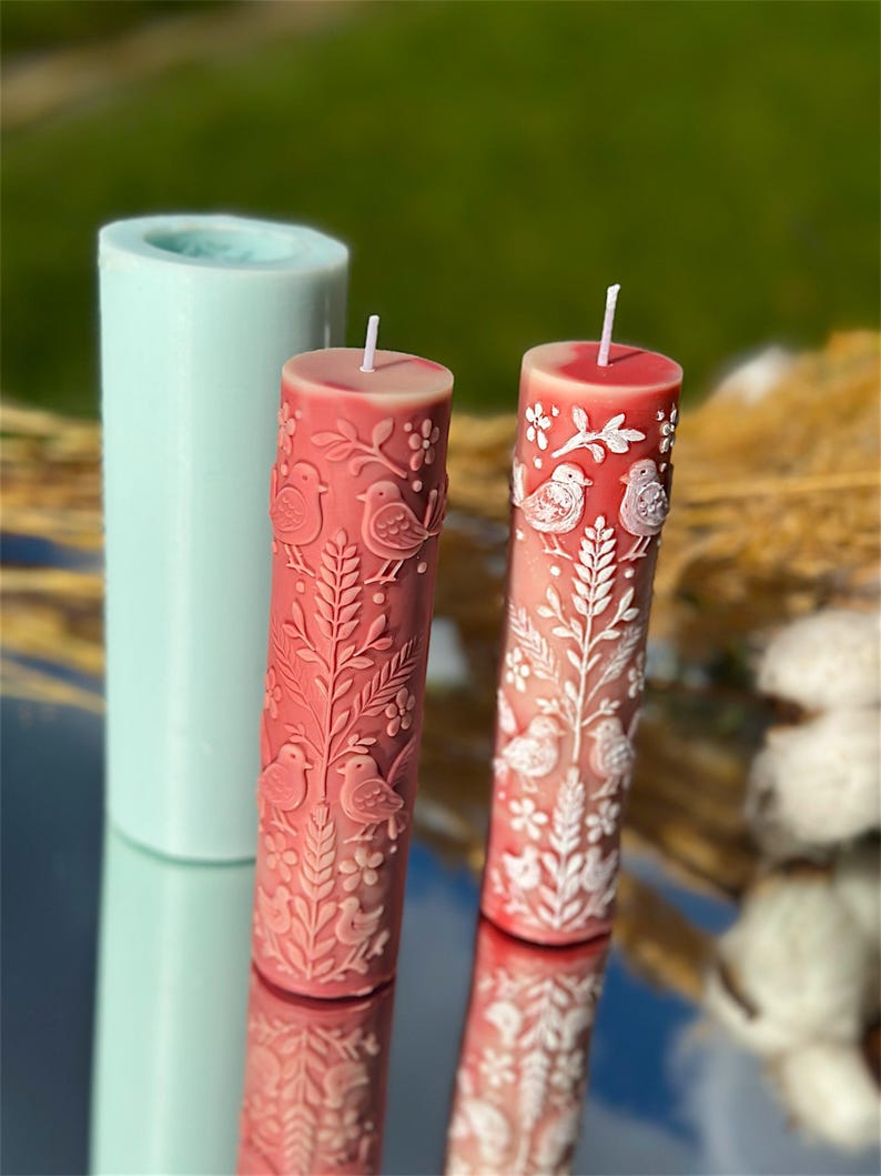 Puede incluir: Dos velas de pilar rojas con dise&ntilde;os de p&aacute;jaros y flores blancos. Un molde cil&iacute;ndrico azul claro para velas est&aacute; en el fondo. Las velas tienen una superficie lisa y reflectante y una mecha blanca.