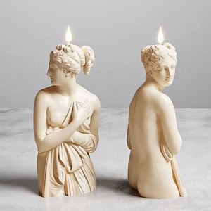 May include: Two off-white candles shaped like classical statues. Each candle depicts a female figure with detailed hair and draped fabric. The flames are lit, creating a unique decorative piece. The candles are set against a neutral background.