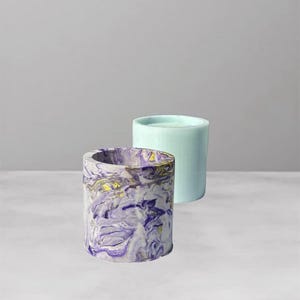 May include: Two cylindrical containers. The front container has a marbled design in shades of purple, white, and gold. The second container is a solid light blue. Both containers are on a white surface.
