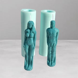 Puede incluir: Dos velas de color turquesa con forma de figuras humanas, una mujer y un hombre, se encuentran frente a dos moldes cilíndricos de velas turquesas. Las velas están detalladas con rasgos anatómicos. El fondo es gris claro.