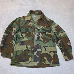 May include: Camouflage pattern jacket with four front pockets and a collared neckline. The jacket features a woodland camouflage design in shades of green, brown, and black. The long-sleeved jacket is made of a durable fabric.