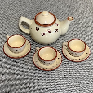 May include: A cream-colored ceramic tea set featuring a teapot and three teacups with saucers. The set is decorated with paw print designs in a dark red color. The rims of the teapot, cups, and saucers are also dark red.