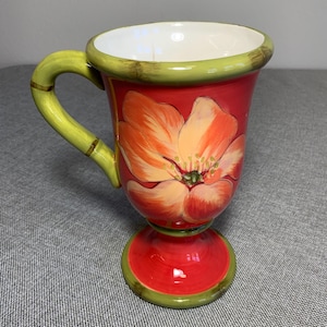 May include: A ceramic mug with a pedestal base and a curved handle, featuring a vibrant red exterior decorated with a large, hand-painted orange and yellow flower. The rim, handle, and base are accented with a muted green trim, and the interior is white.