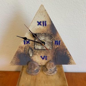 May include: A triangular ceramic clock with a beige and blue color scheme. The clock face features Roman numerals and a square cutout. The clock hands are black, and the hour markers are blue. The clock rests on a wooden base with two decorative ceramic pieces.