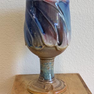 以下が含まれることがあります： 青、紫、茶色の色合いの渦巻き模様が施された手作りのセラミックゴブレット。ゴブレットは、広いボウル、細いステム、詳細なベースを備えています。木製の表面に展示されています。