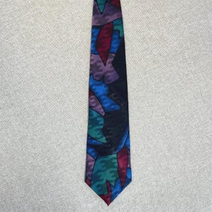 May include: A necktie featuring an abstract pattern of overlapping shapes in shades of deep blue, purple, teal, and dark red. The design consists of irregular, jagged forms that create a layered, multi-colored appearance across the entire fabric surface.