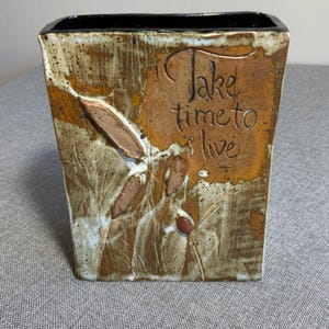 Op de afbeelding: Een rechthoekige keramische vaas met een gespikkeld bruin en beige glazuur. De voorzijde is voorzien van een reliëf met botanisch ontwerp en de handgekerfde tekst "Take time to live" in een schrijflettertype. De binnenrand is zwart geglazuurd.