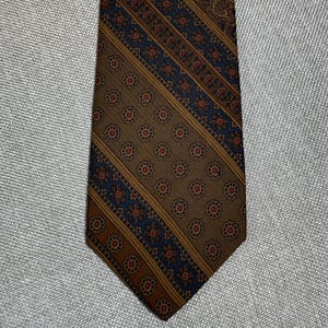 May include: A necktie featuring a repeating pattern of small floral motifs in red and white. The design is divided into diagonal sections of brown and dark blue, with thin gold-colored lines separating the distinct patterned bands throughout.