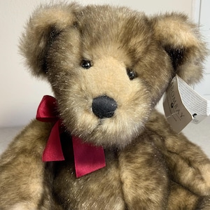 Könnte beinhalten: Ein Teddybär mit braunem und beigem Fell, einer schwarzen Nase und einer roten Schleife um den Hals. Der Bär hat ein Etikett an seinem Ohr. Ein klassisches Stofftier.